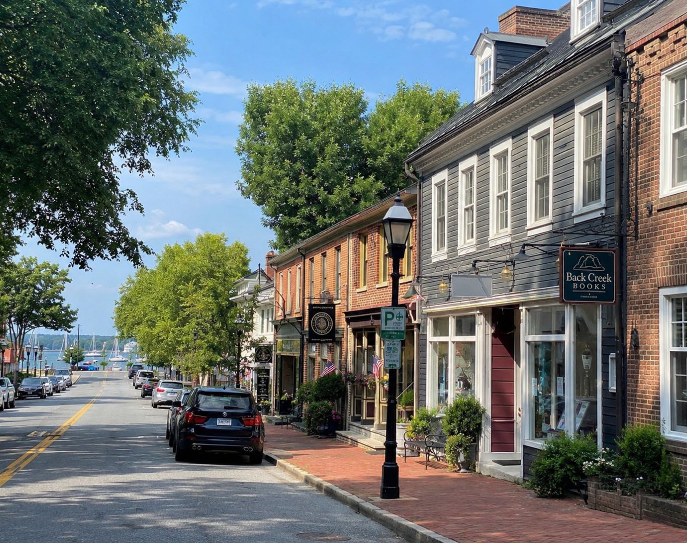 West Annapolis, MD street scene with historic storefronts, brick sidewalks, parked cars, trees, and a waterfront view in the distance